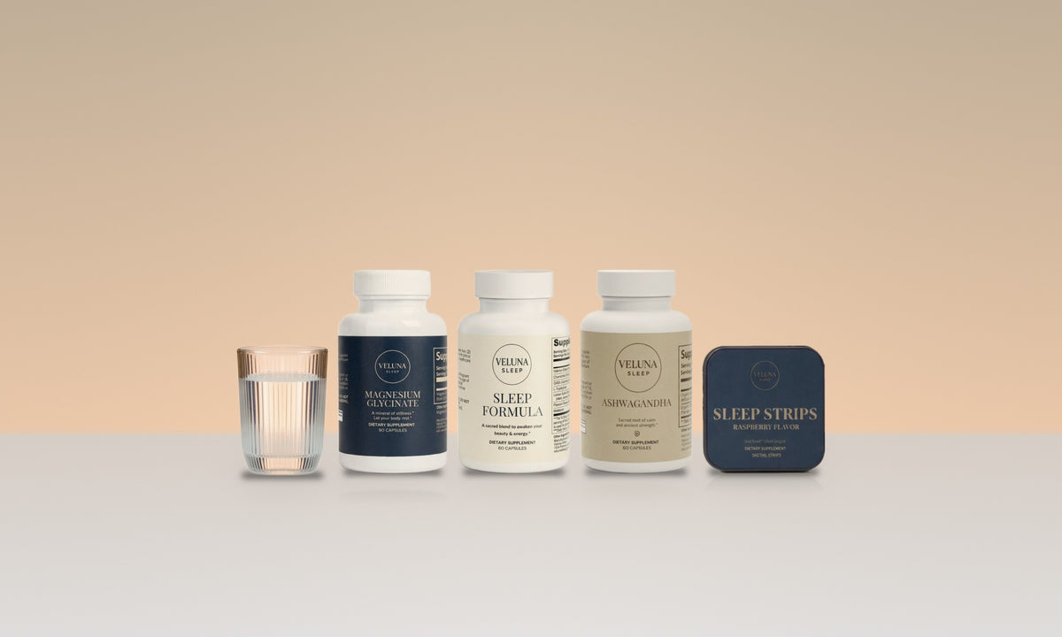 The four Veluna Mind Calm products, Magnesium Glycinate, Sleep Formula, Ashwagandha and Sleep Strips, in a row beside a glass of water on a warm studio backdrop.