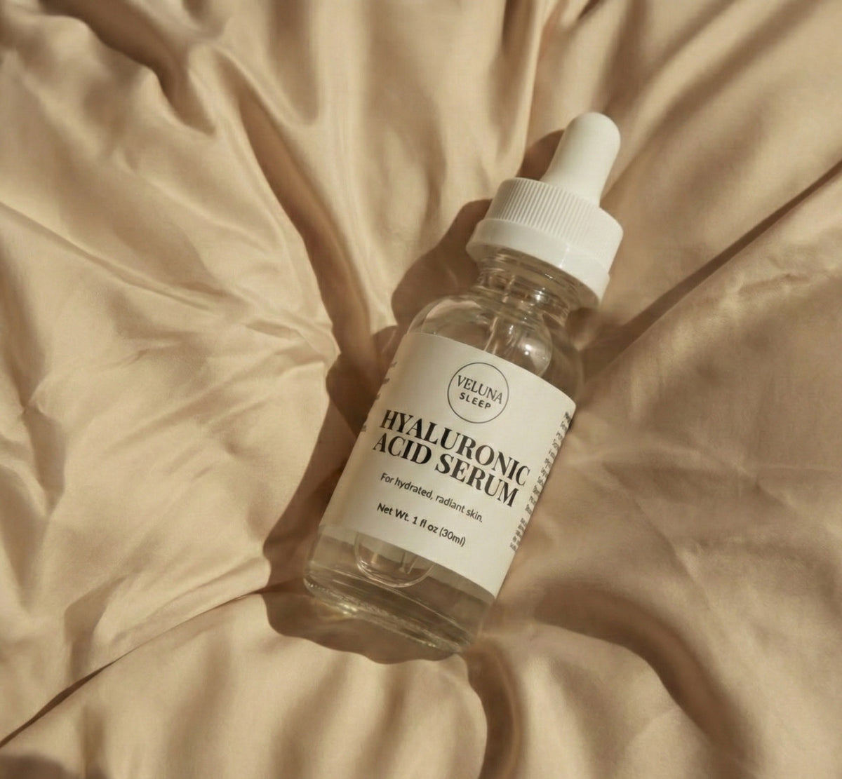 Veluna Hyaluronic Acid Serum dropper bottle lying on sunlit, rumpled cream bedding, seen from above