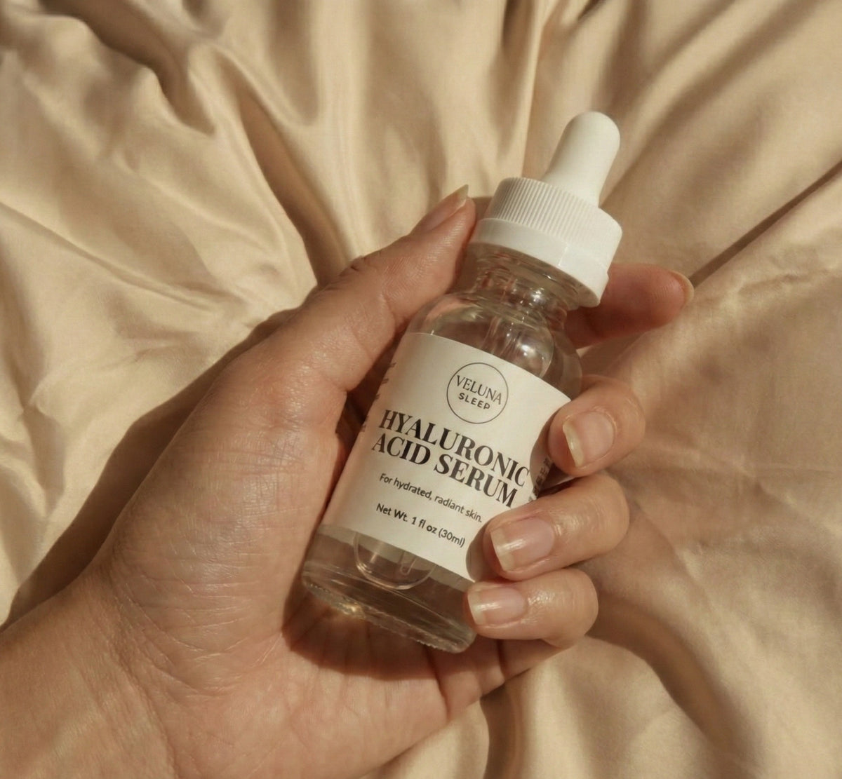 Veluna Hyaluronic Acid Serum dropper bottle cradled in one hand over cream satin sheets