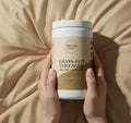 Veluna Grass-Fed Collagen Peptides, Chocolate, tub held upright in both hands over cream satin bedding