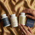 Veluna Mind Calm Full Bundle laid flat on golden satin, two hands with gold rings beside the Ashwagandha bottle